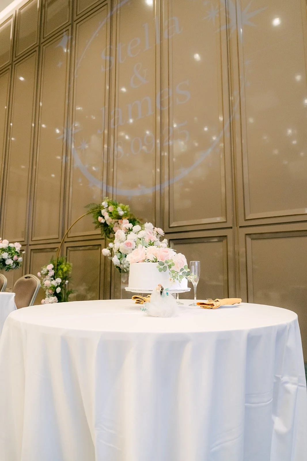 The white wedding cake on a round table in front of the ballroom wall