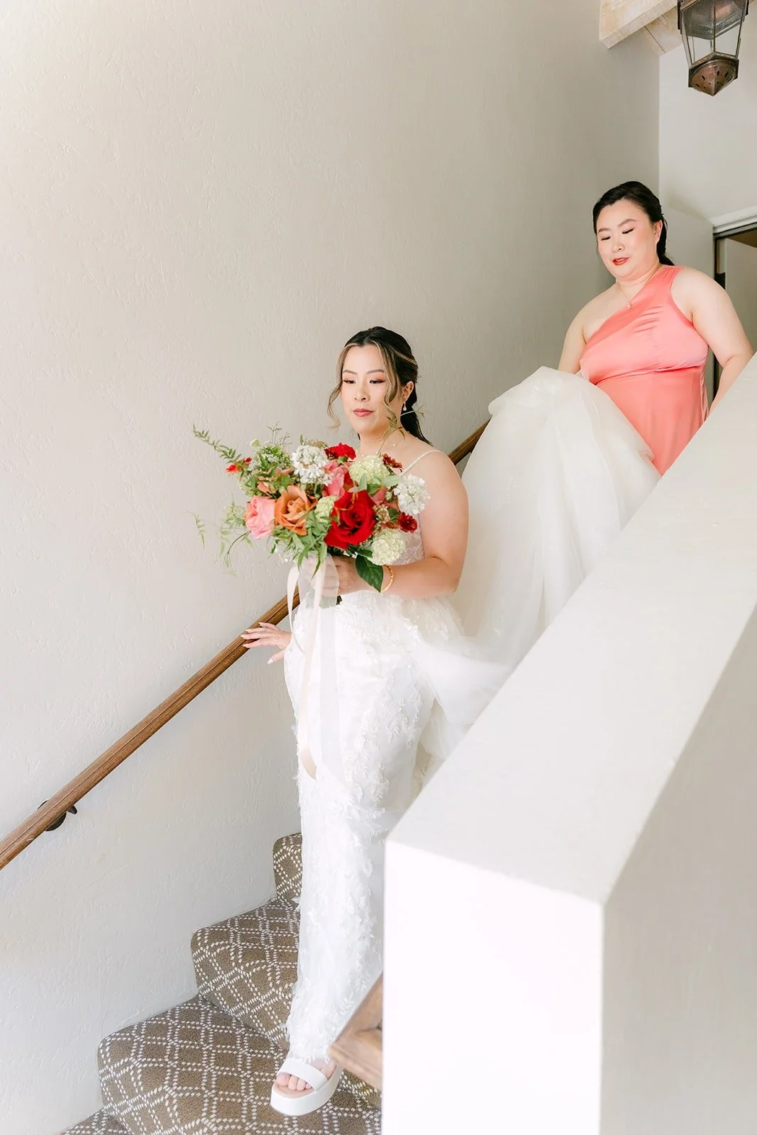 Joanne walking down the stairs with her bouquet, a bridesmaid helping with her train