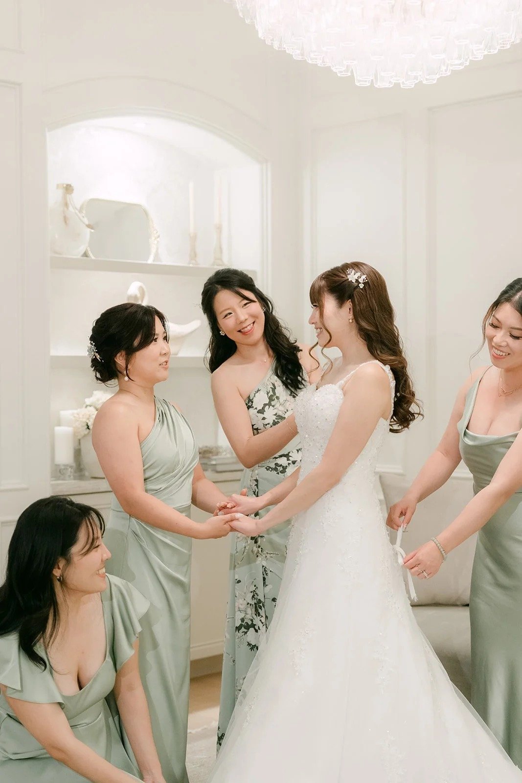 Bridesmaids in sage green holding hands with Marina as she finishes getting ready