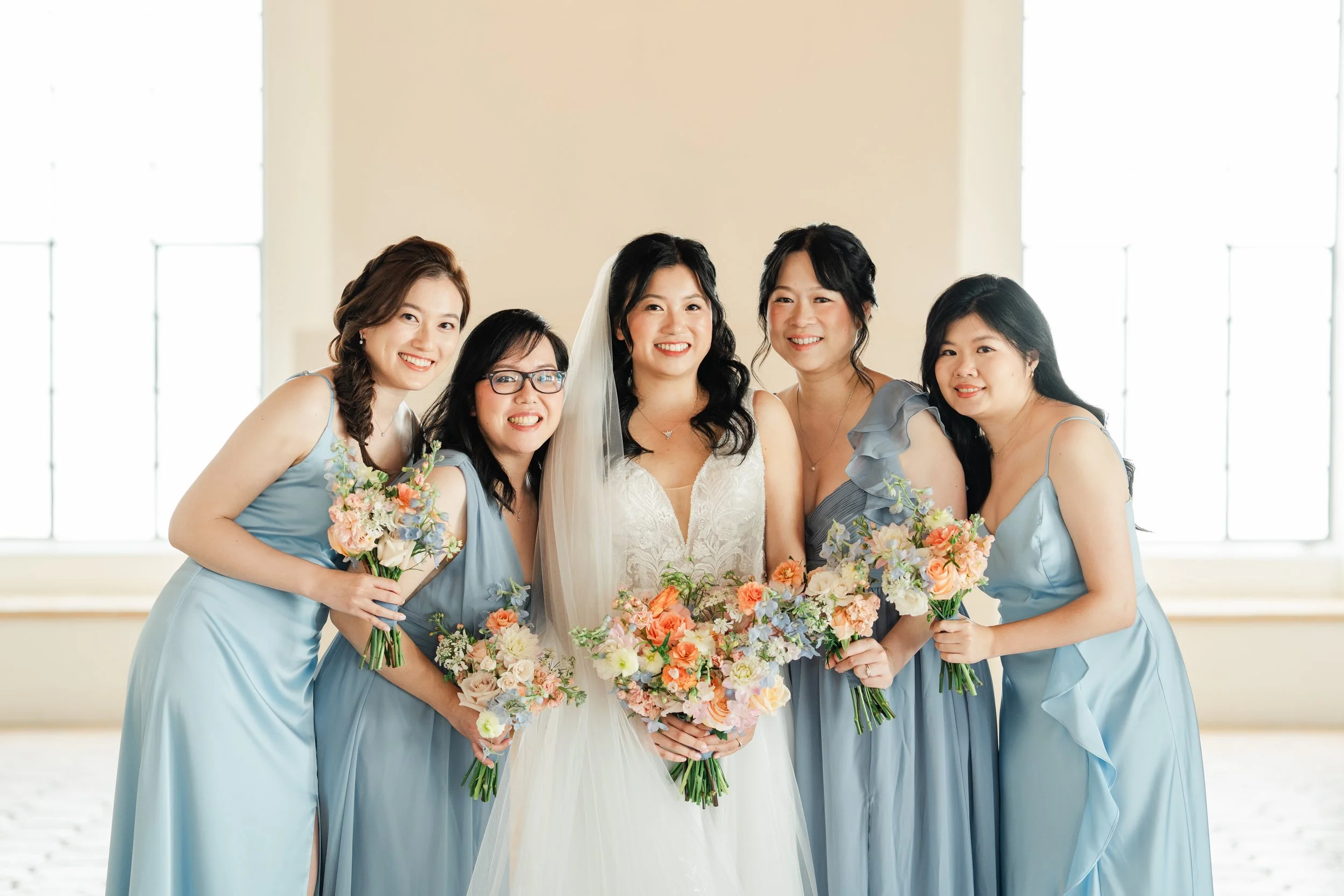 A bride in a white wedding gown and veil standing among five women in light blue dresses holding bouquets of flowers, inside a room with large windows.