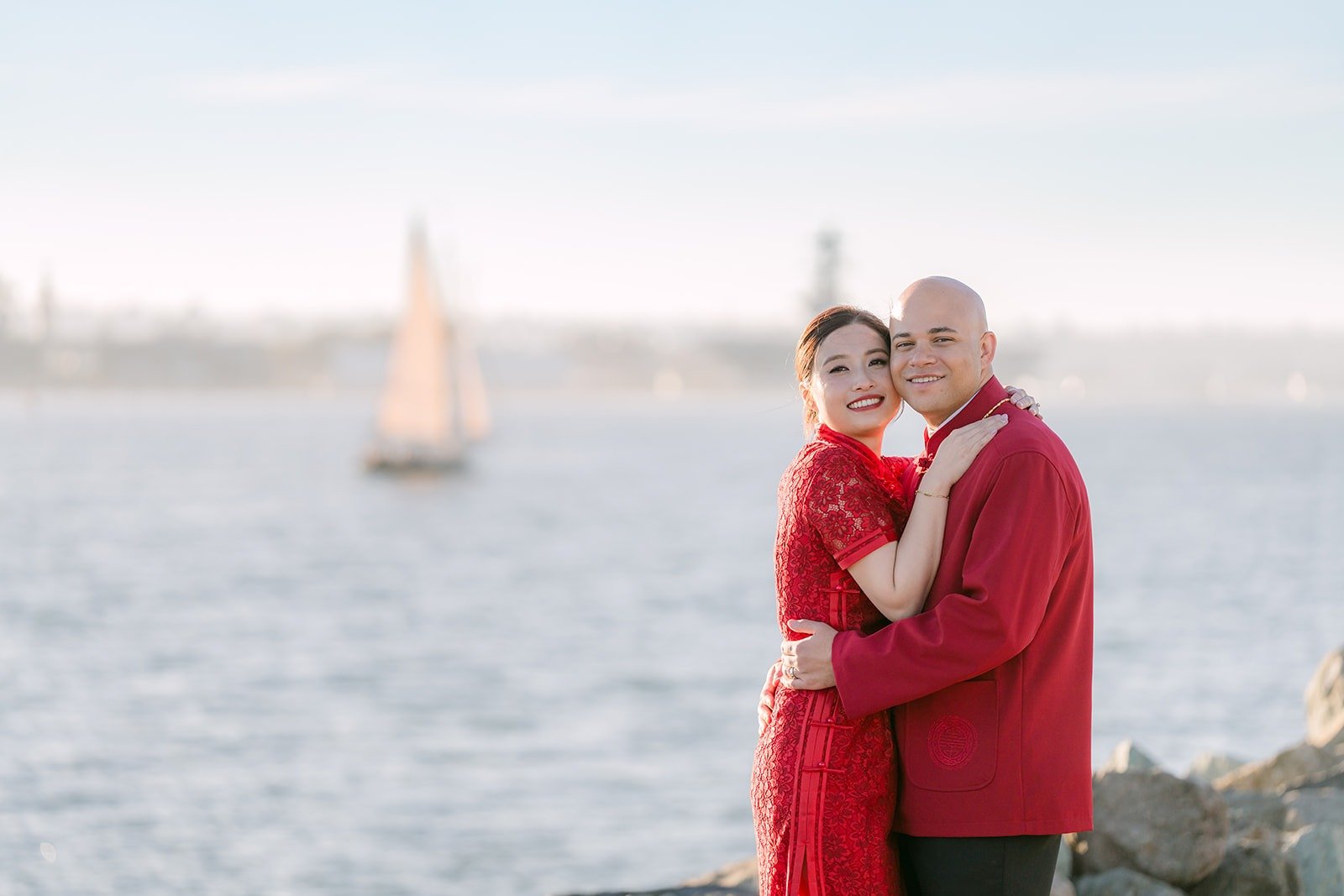 Elisa and Brandon embrace on the pier with a sailboat behind them