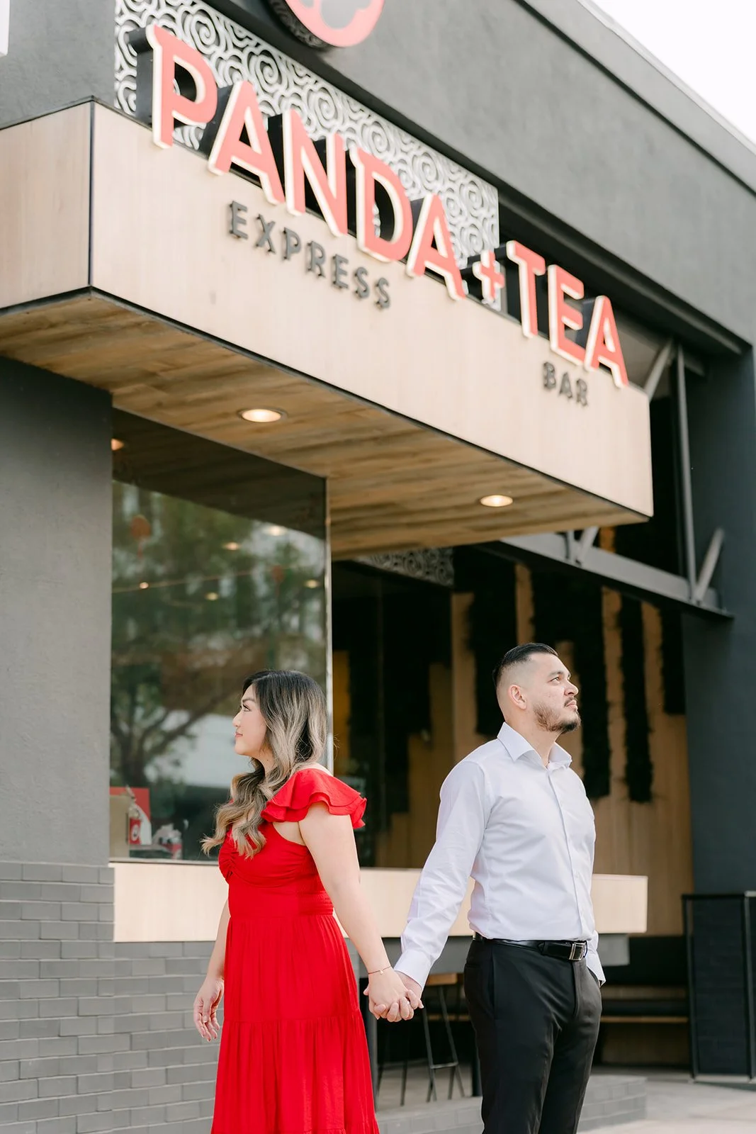Linh in a red dress and Ankush walk hand in hand beneath the Panda Express sign.