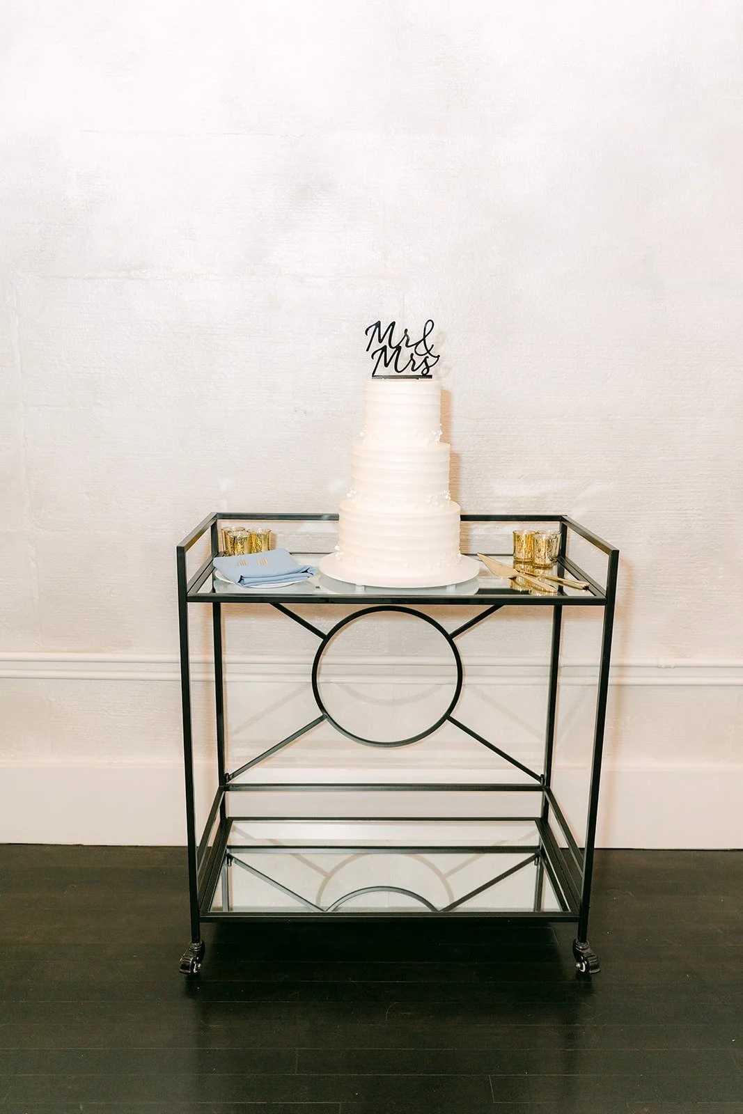 The white tiered cake on a gold bar cart in the reception room, photographed by One and Only Production