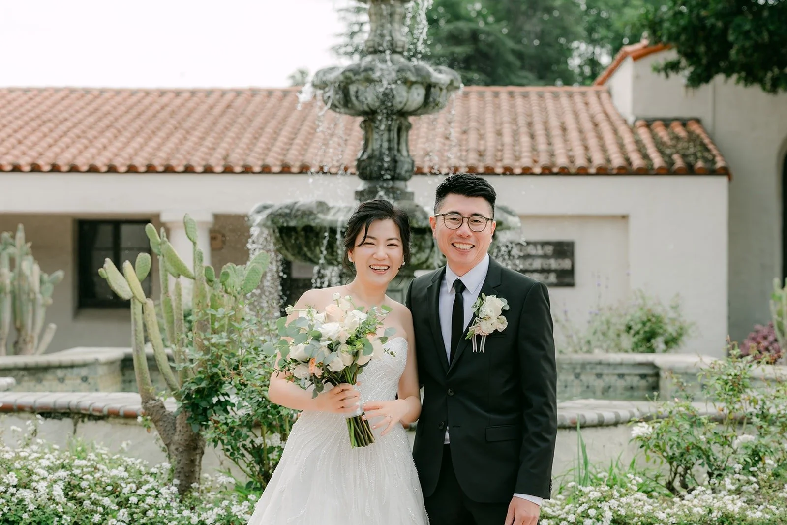 Eunice and Eric smile together in front of the fountain and red tile roofs at San Gabriel Mission