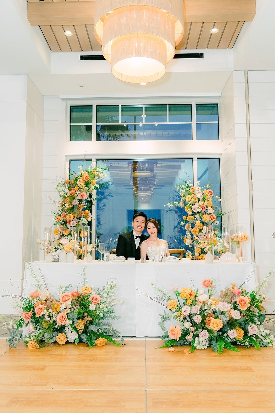 Angela and Eli at their sweetheart table framed by peach florals in the ballroom