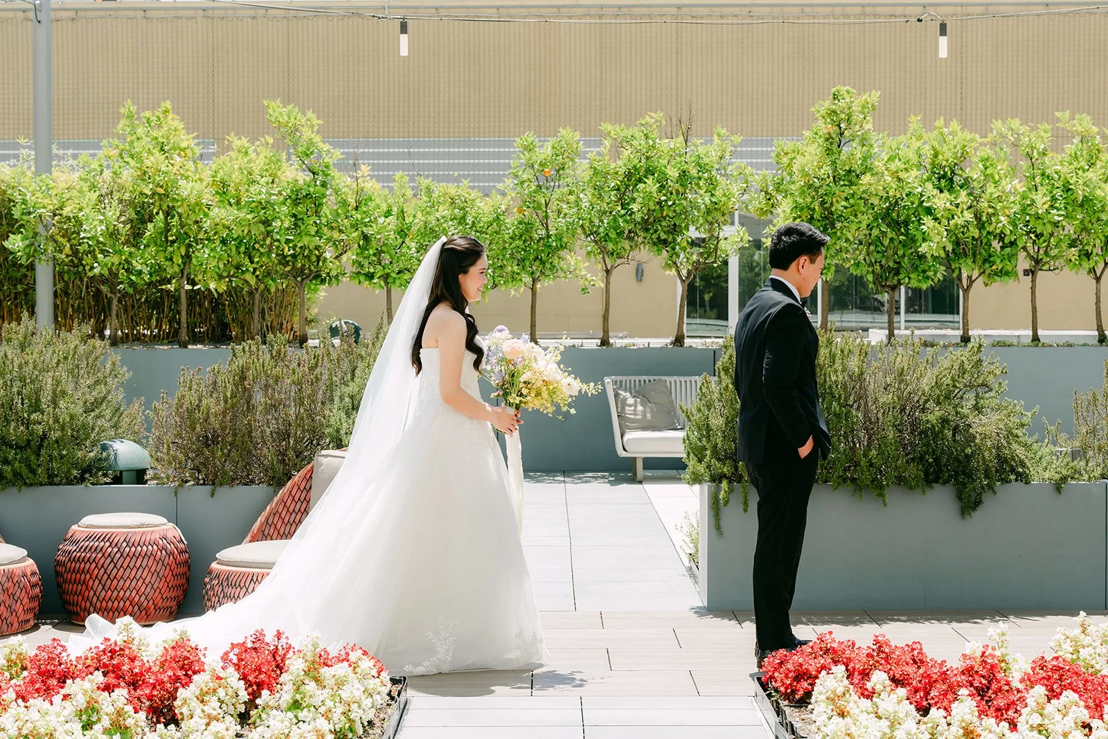 Bride walking up behind Hoan for their first look on the terrace among red flowers