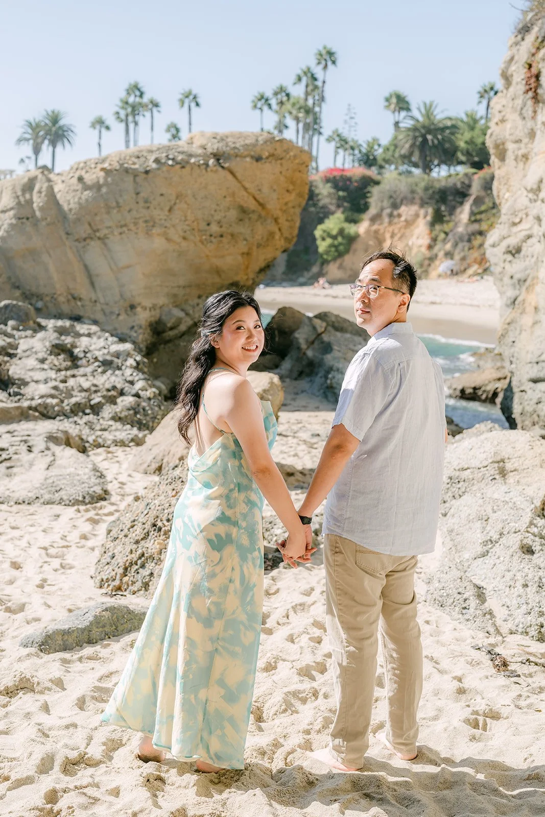 Constance and Lawrence holding hands by the rocks with palm trees above.