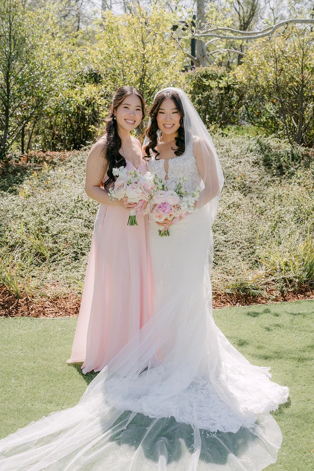 Kelli and a bridesmaid smiling together on the lawn with her train spread across the grass