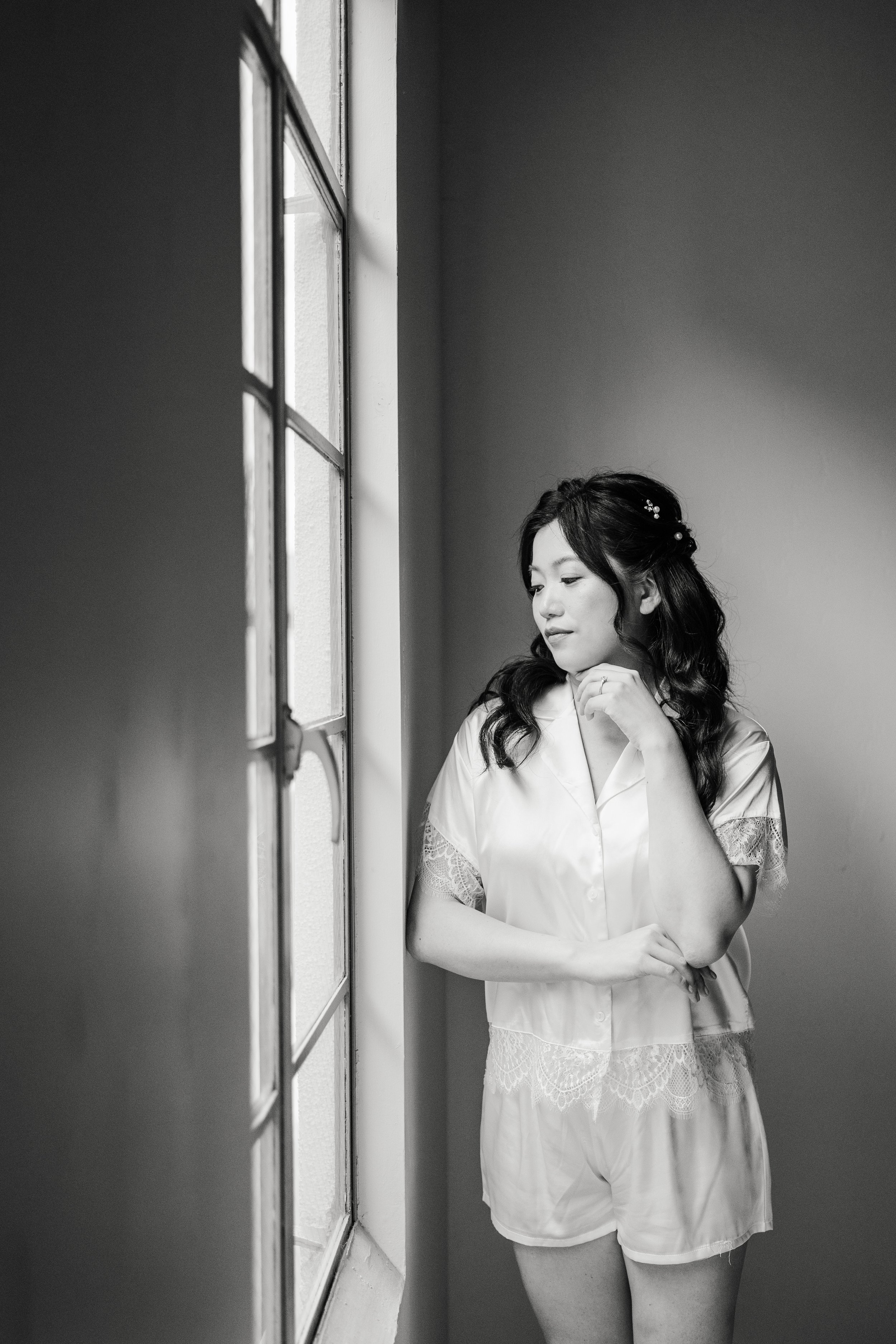 A woman in satin and lace pajamas standing by a window, looking down thoughtfully, with her hand near her chin, in a black-and-white photo.