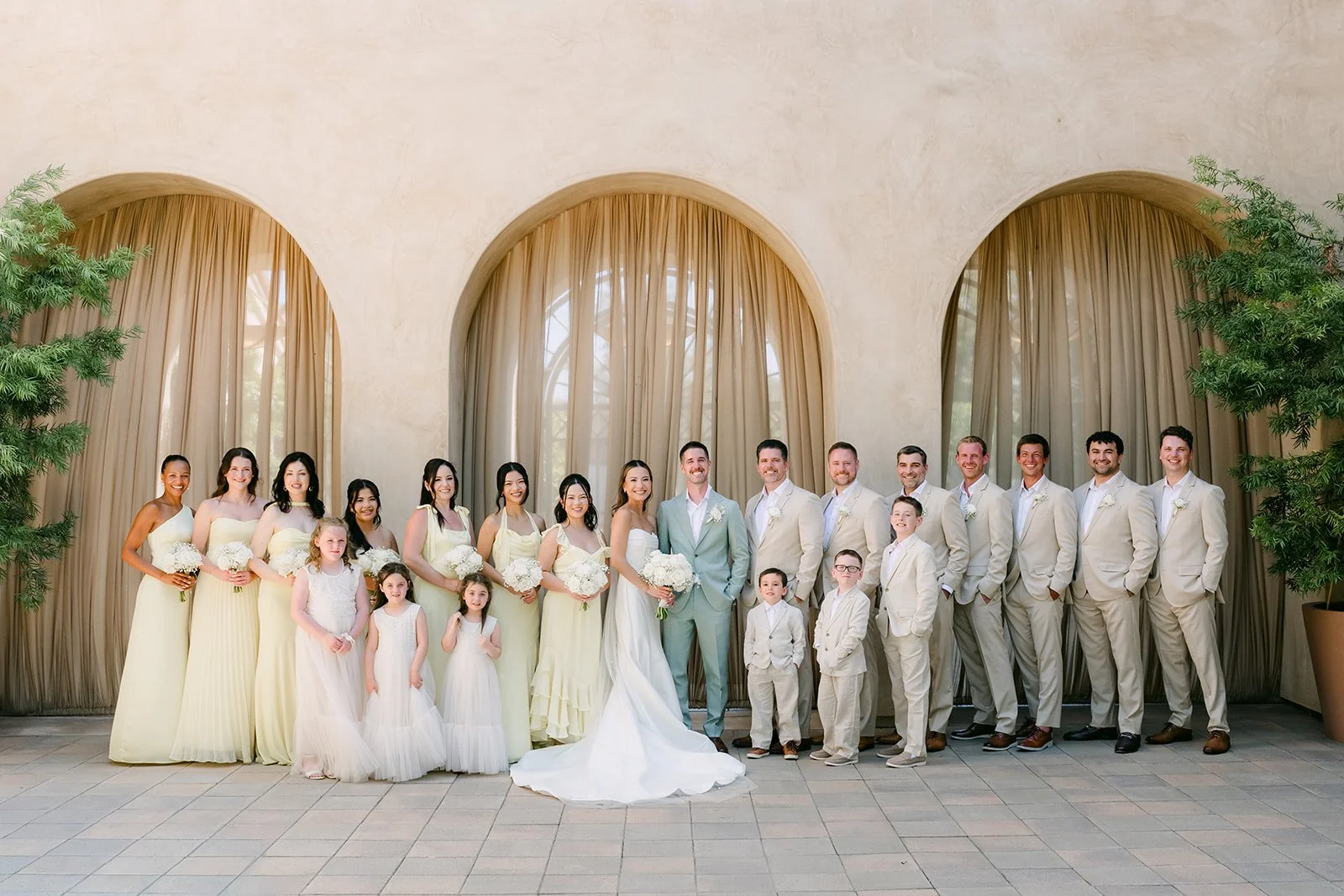 Full wedding party in butter yellow and sand linen lined up beneath the arches