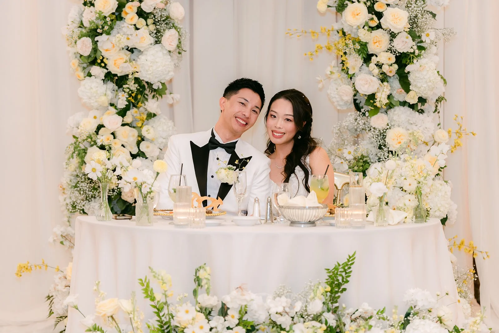 Jenny and Ryan laughing at their sweetheart table with white florals, photographed by One and Only Production.