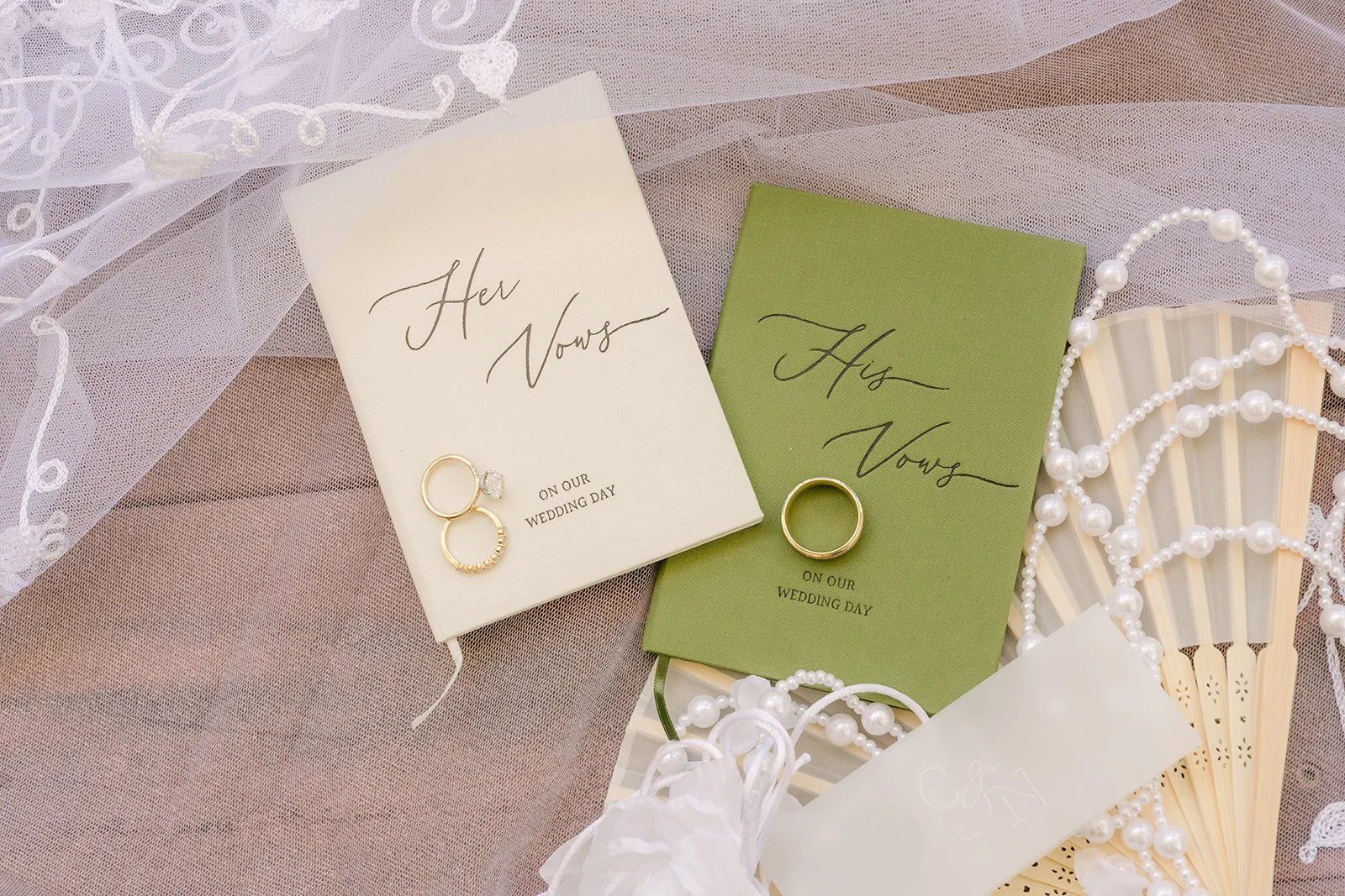 Ivory and pistachio green His Vows and Her Vows books with wedding rings, a veil and a gold fan in Nicole's flat lay.