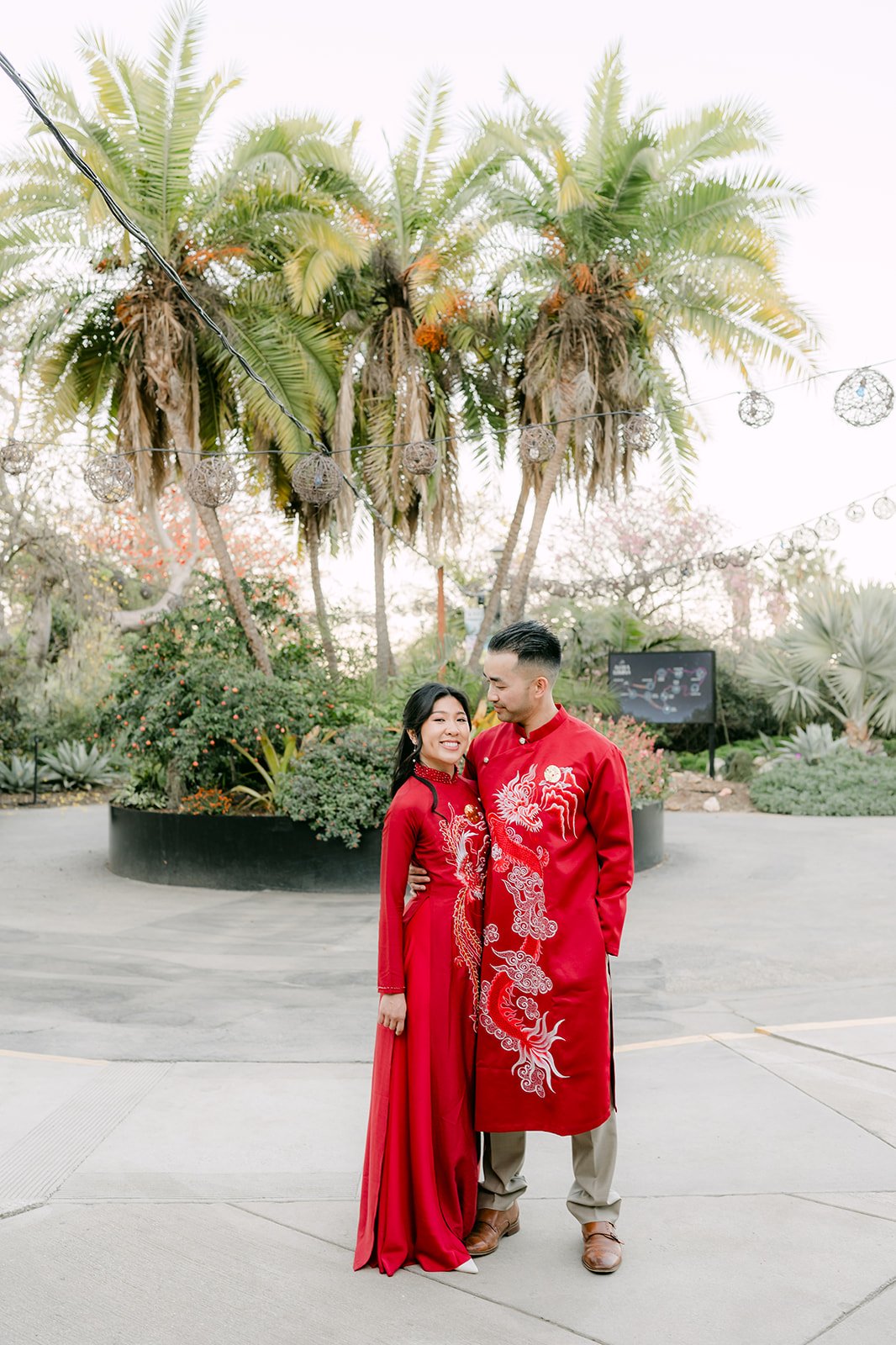 Kim and Vincent in matching red ao dai in a palm-lined courtyard.