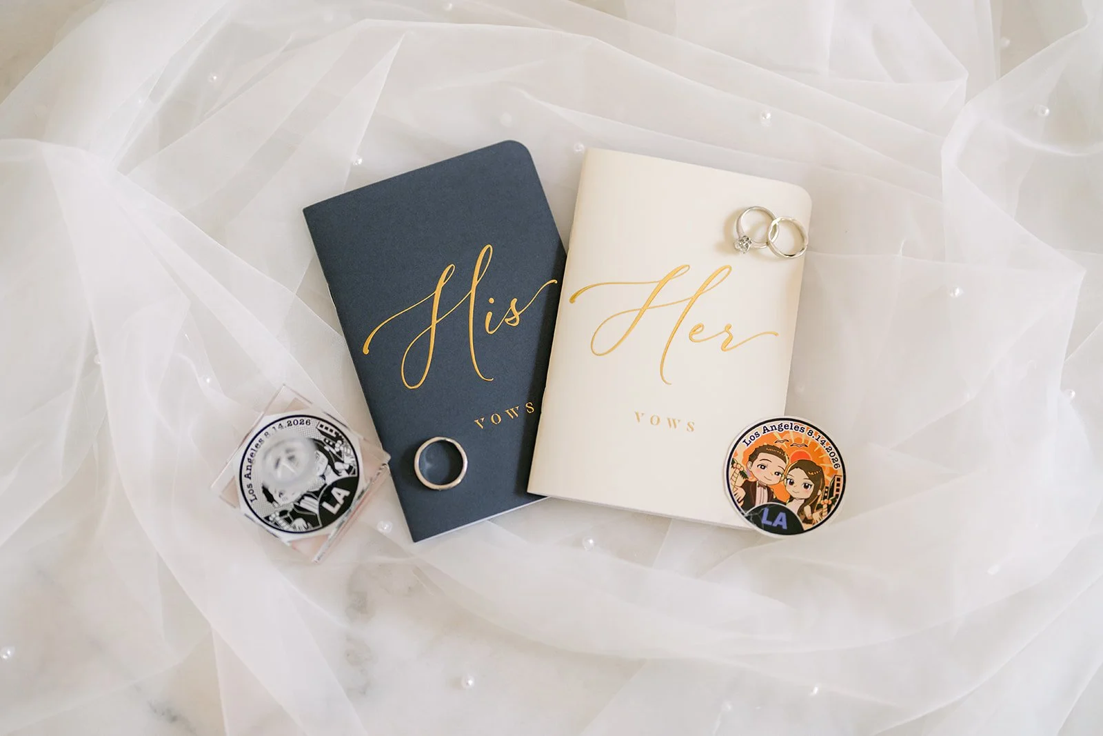 His and her vow books with wedding bands and couple pins on tulle and marble