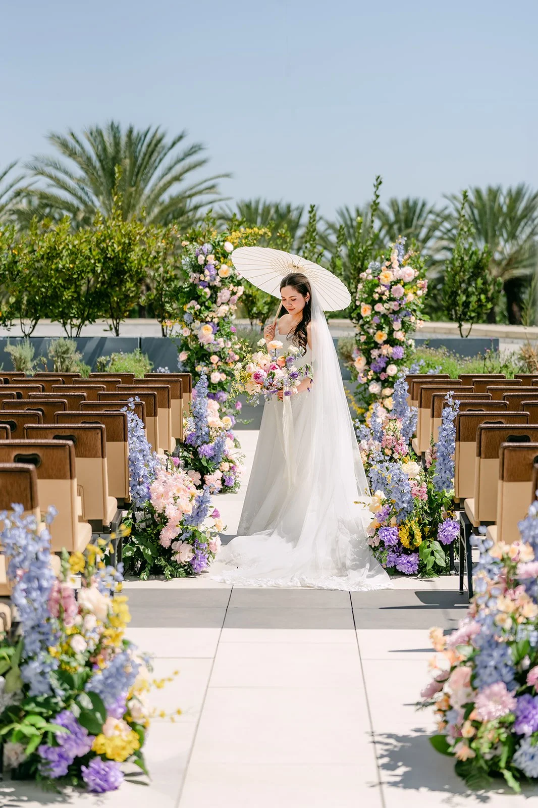Bride holding a white parasol at the end of the flower-lined ceremony aisle