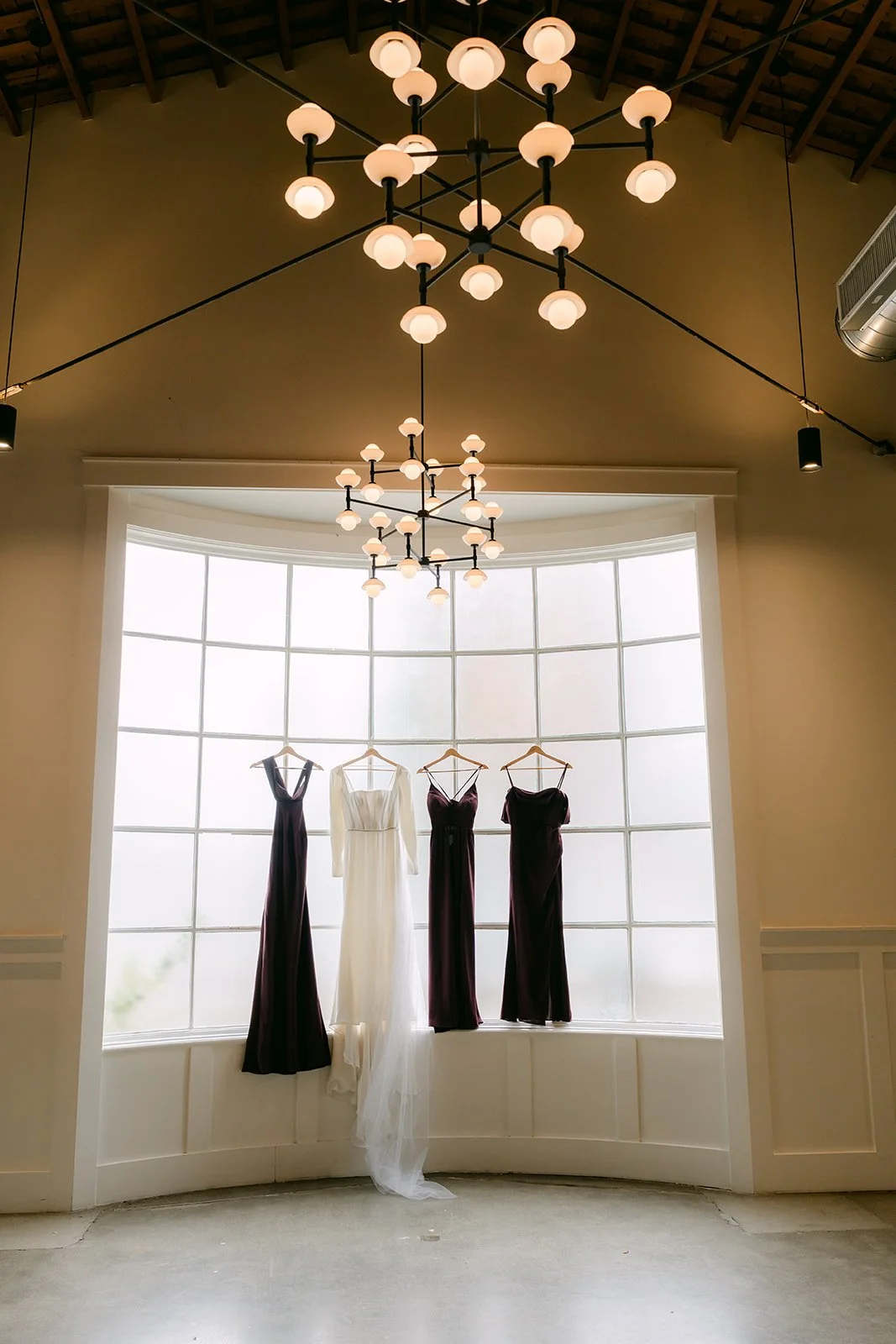 Bridesmaid dresses and Denise's gown hanging in the tall window under a chandelier at The Richland.
