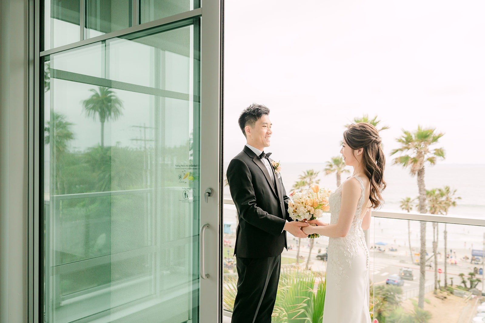 Angela and Eli facing each other on the balcony, palms and ocean beyond