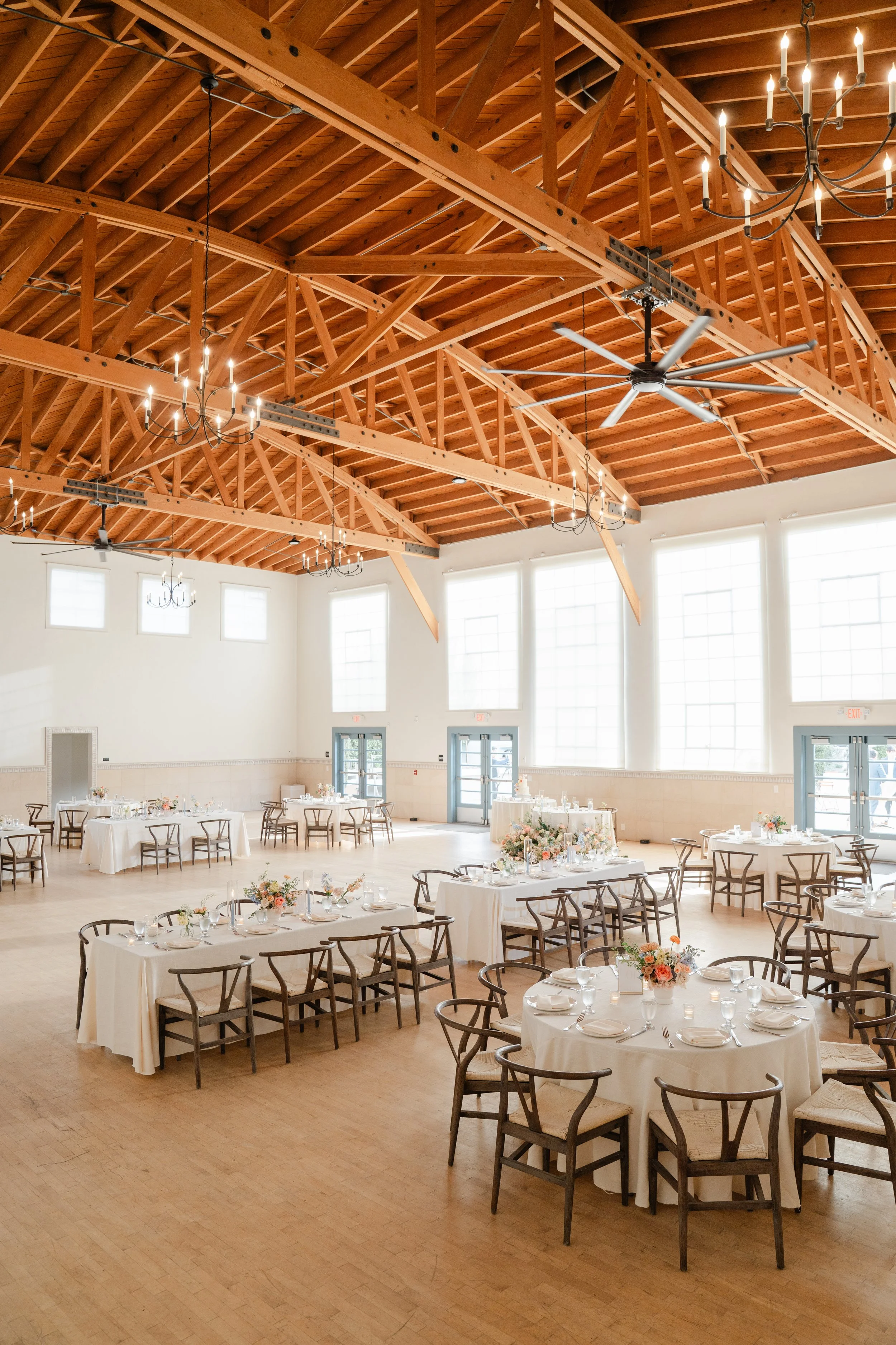 A spacious banquet hall with high wooden ceiling beams, large windows, and decorated round tables with white tablecloths, floral centerpieces, and table settings, ready for an event.