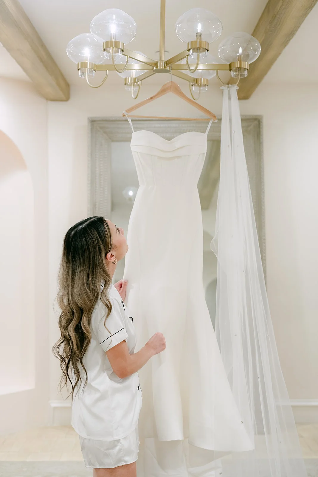 Bride with long waves admiring her wedding gown hanging under a gold chandelier