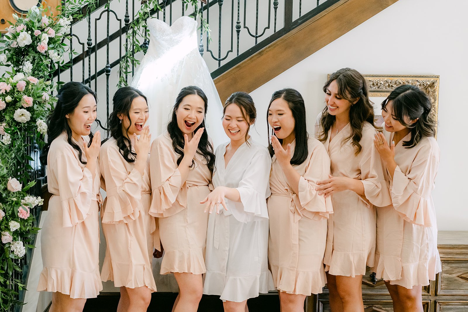 Bridesmaids in blush robes laughing with the bride by the floral staircase