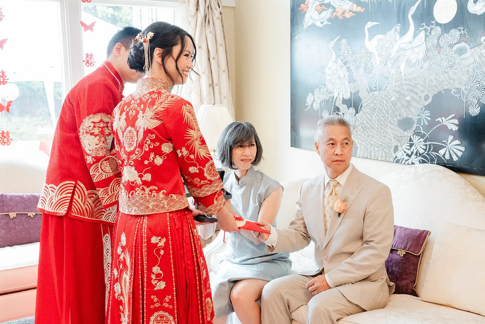 A bride and groom in red qun kwa serving tea to their parents on the sofa.