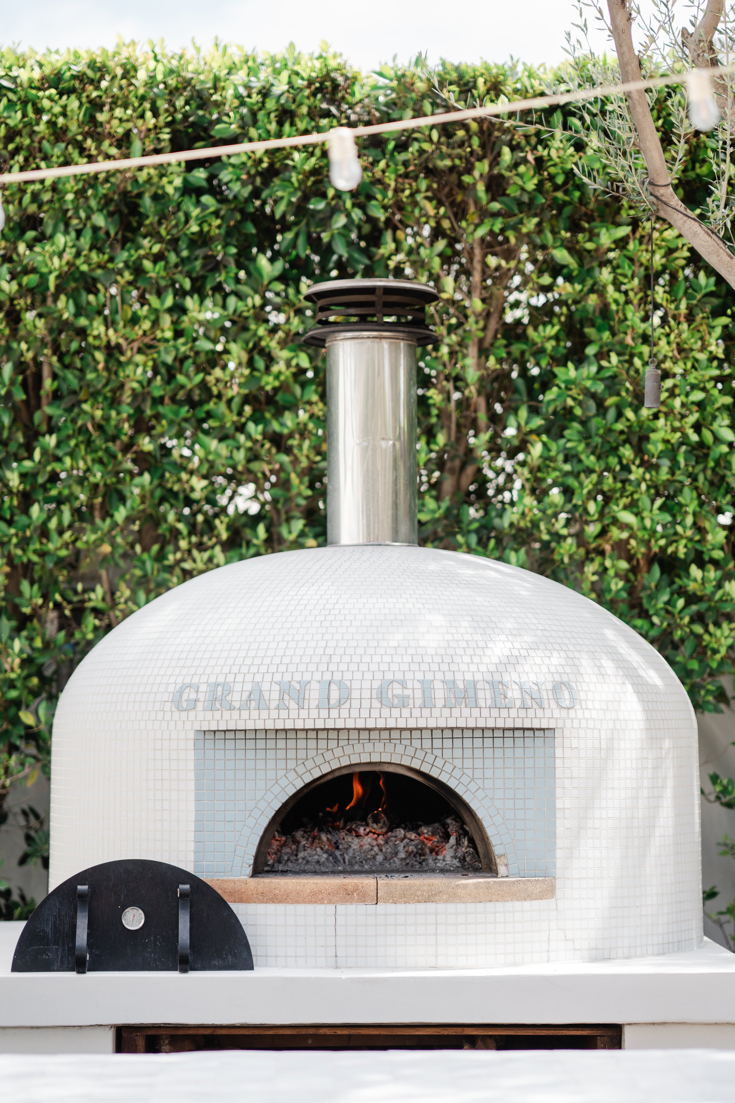 An outdoor pizza oven with a rounded white tiled exterior, a small black charred arch, and the words 'GRAND GUVEN' on top, with a chimney on top of the oven. There are green bushes behind it and a string of outdoor lights hanging above.