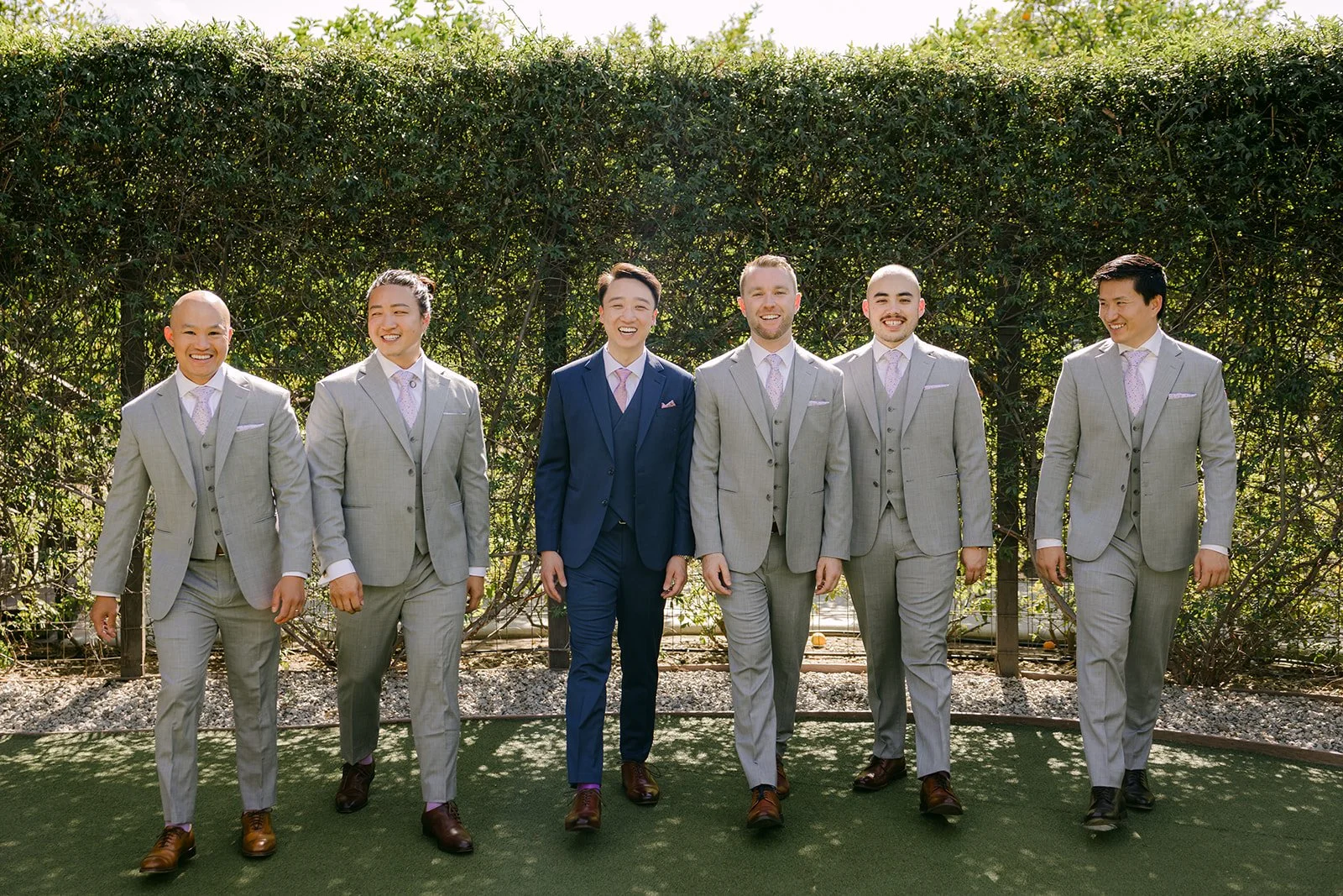 Elijah in navy with his groomsmen in gray in front of a hedge wall.