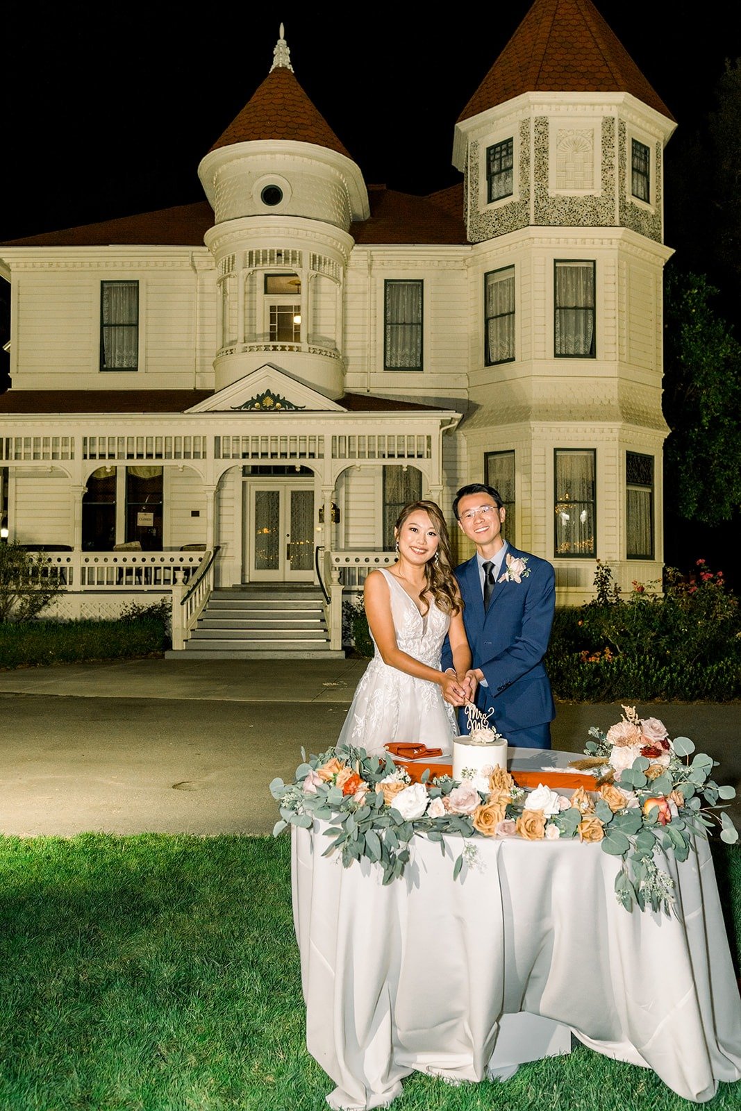 Ariel and Alex beside their cake in front of the lit ranch house