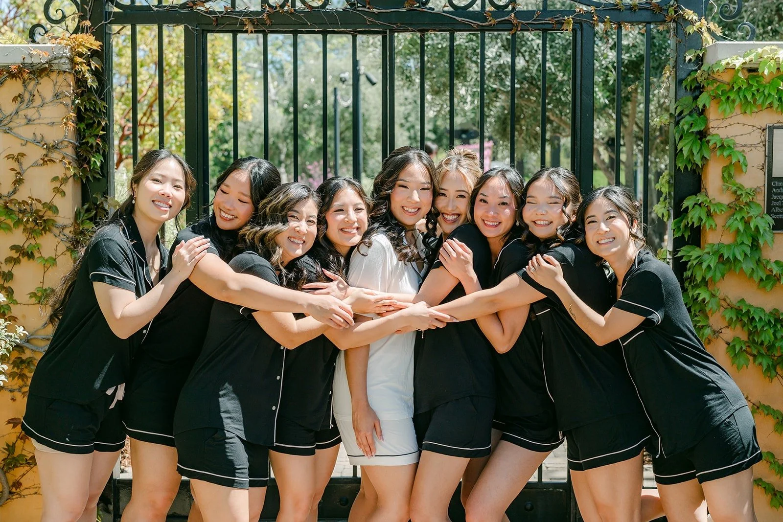 Kelli and her bridesmaids in black satin pajamas laughing in front of an iron gate at Los Robles Greens
