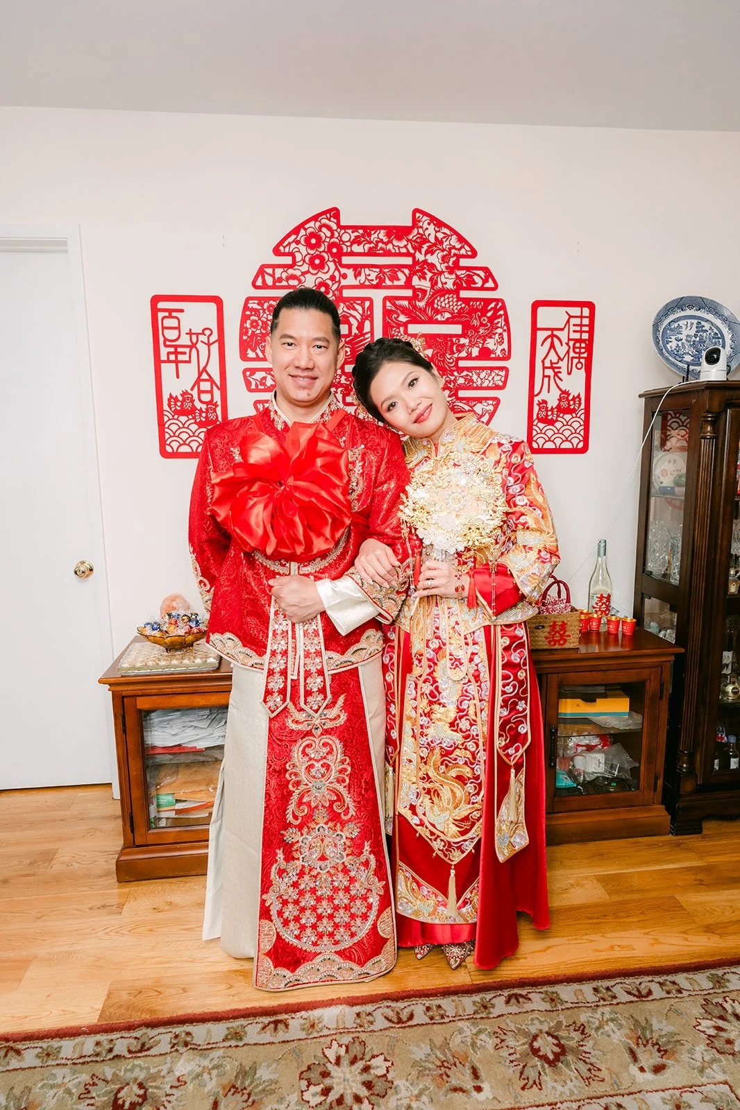 A bride and groom in red traditional attire posing in the living room.