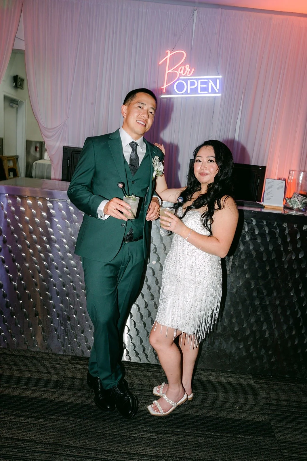 Jennifer and Kevin toast at the bar under a bar open neon sign