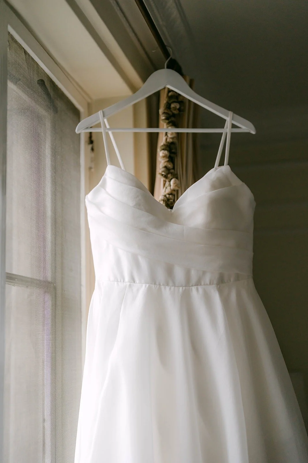 Saira's sweetheart neckline wedding dress hanging on a wooden hanger by the window