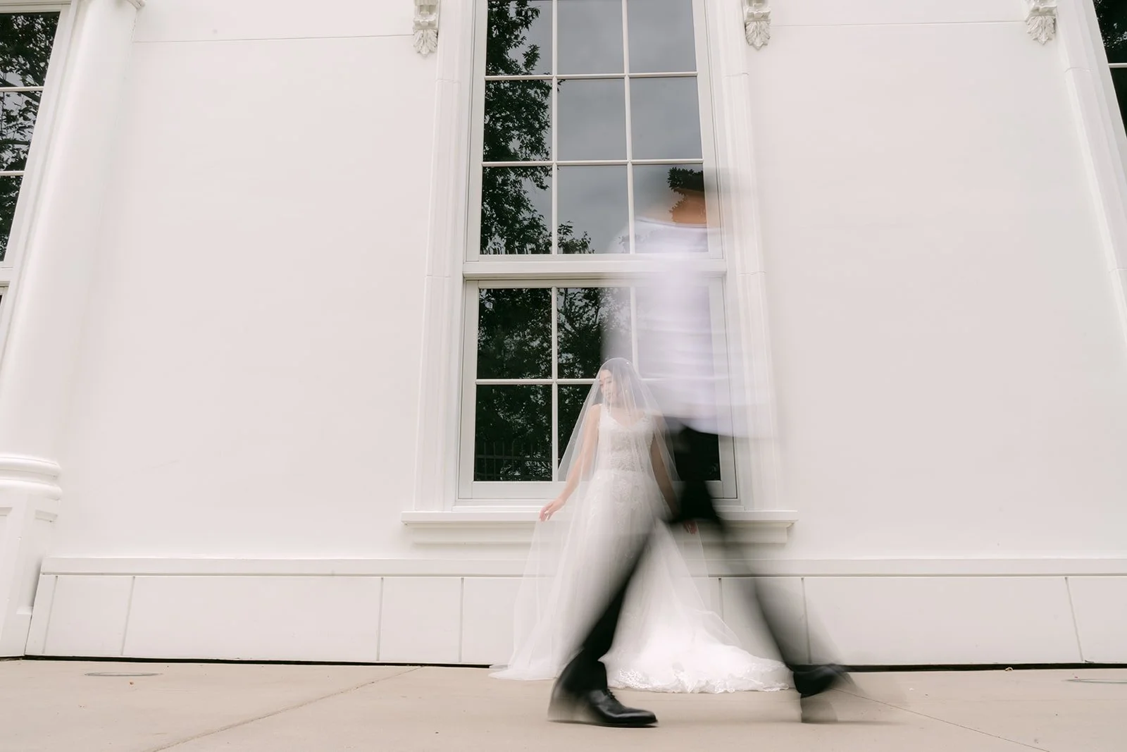Motion blur portrait of Jenny and Ryan walking past a white building.