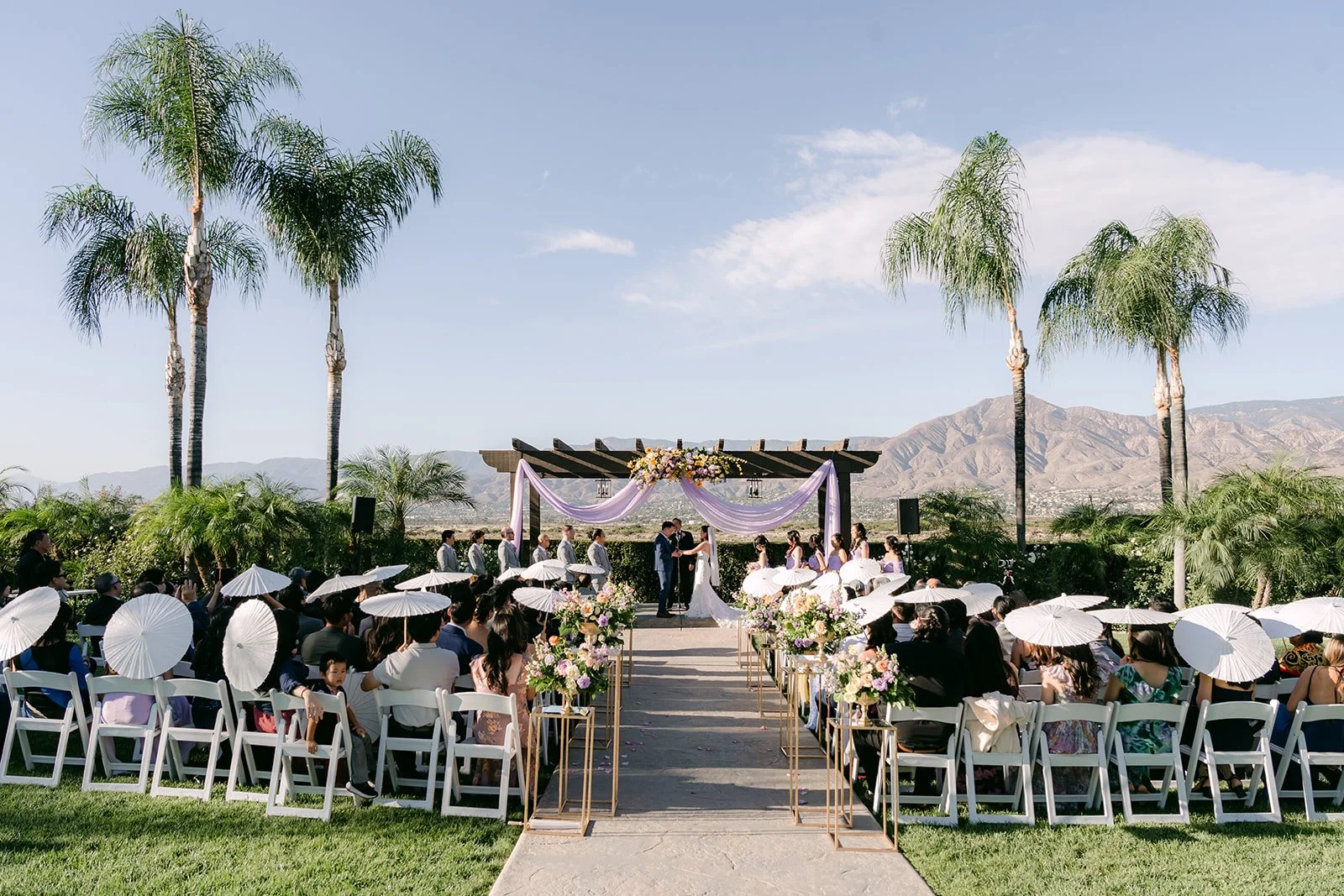 Wide view of the ceremony with palm trees, white parasols and mountains.