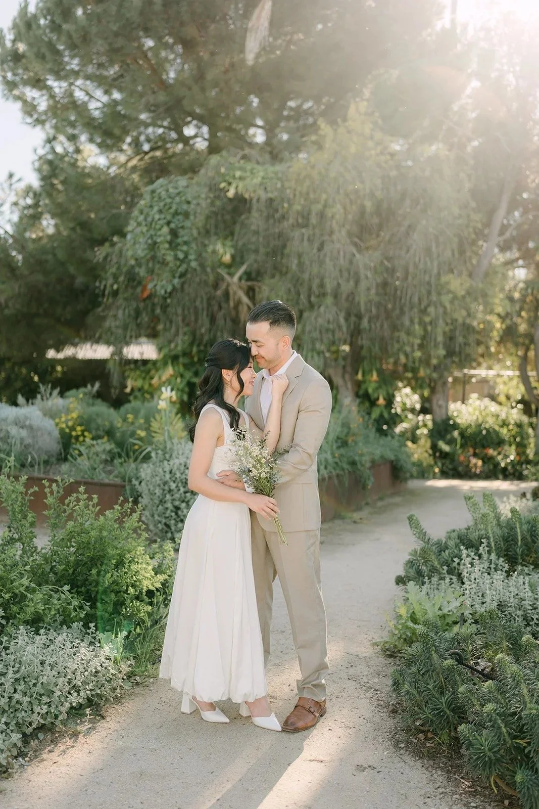 Kim in white and Vincent in a tan suit walking hand in hand down a sunlit garden path.