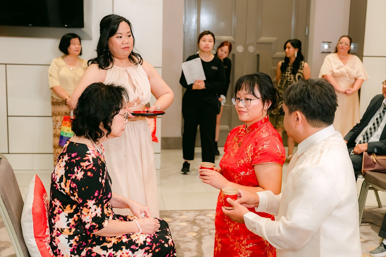 Stella in her red qipao serving tea to a family member as guests watch