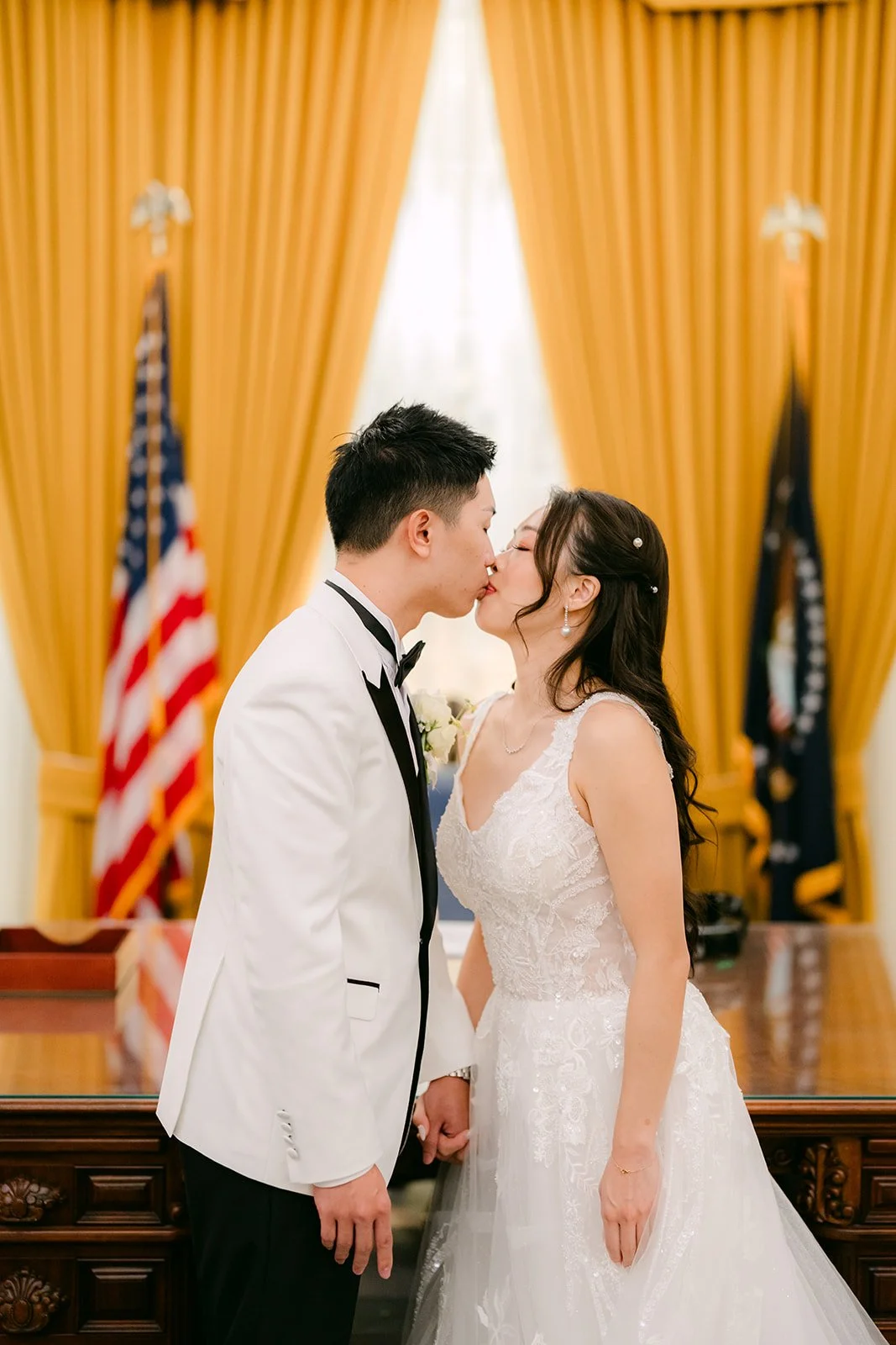 Jenny and Ryan kissing in front of the flags and gold curtains of the Oval Office replica.