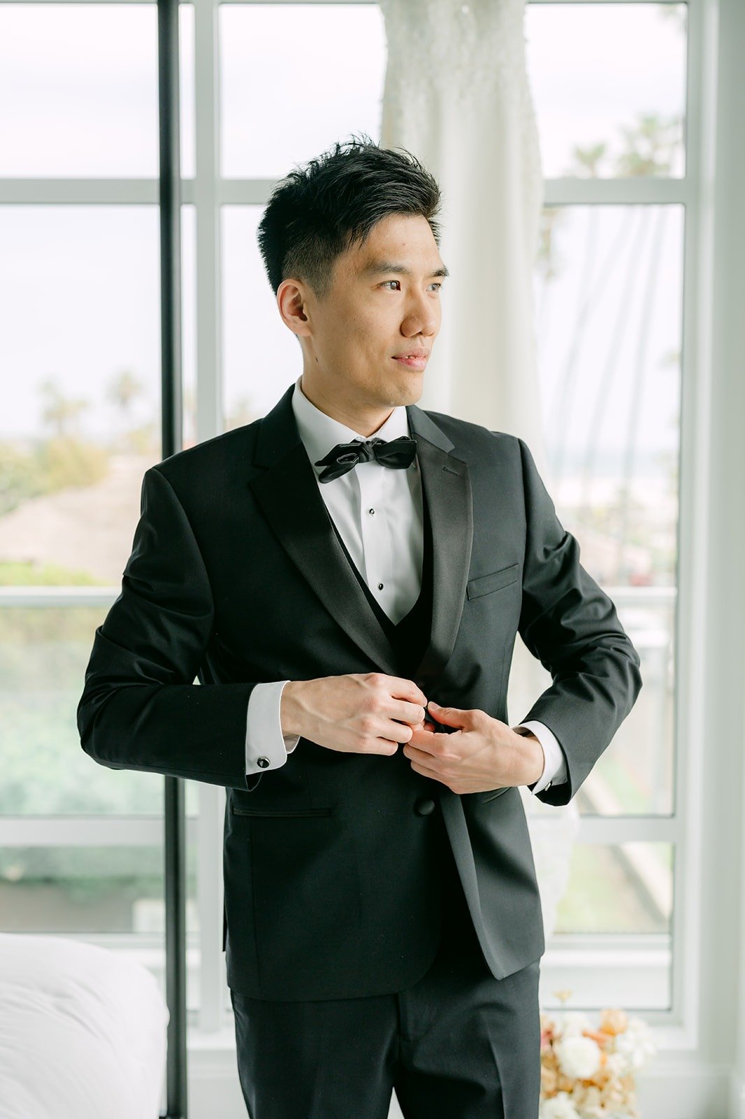 Eli buttoning his black tuxedo jacket in the window light