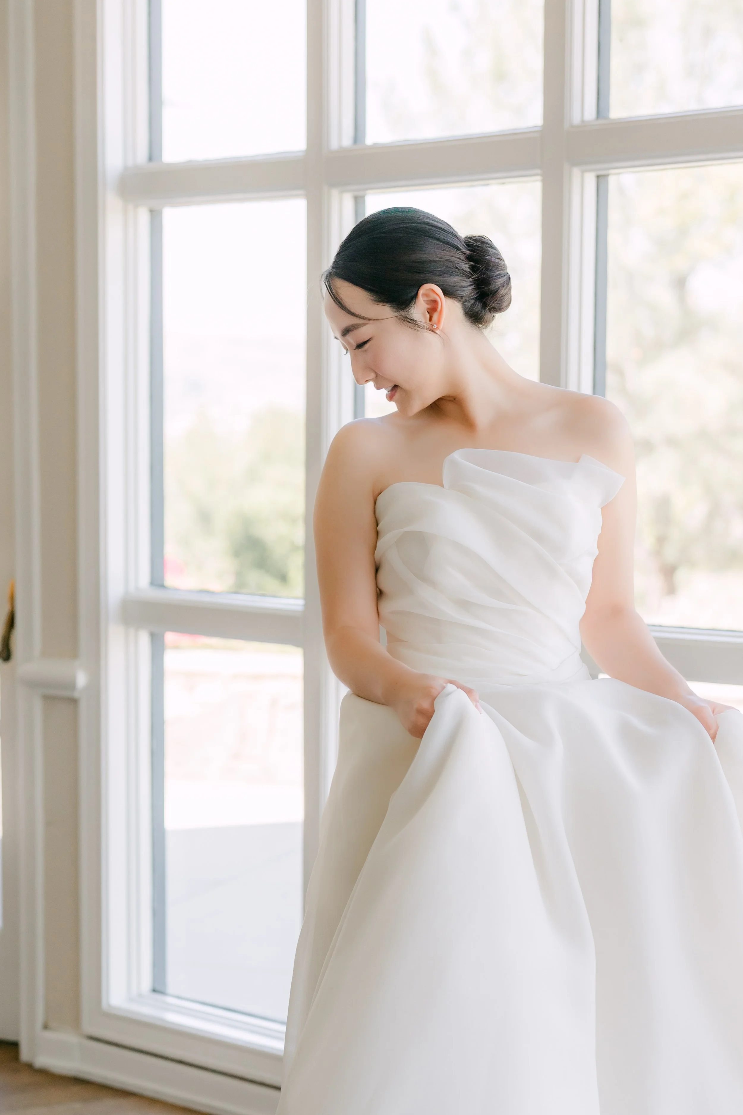 A young woman in a strapless white wedding dress sitting by a large window with natural light, smiling gently and looking down.