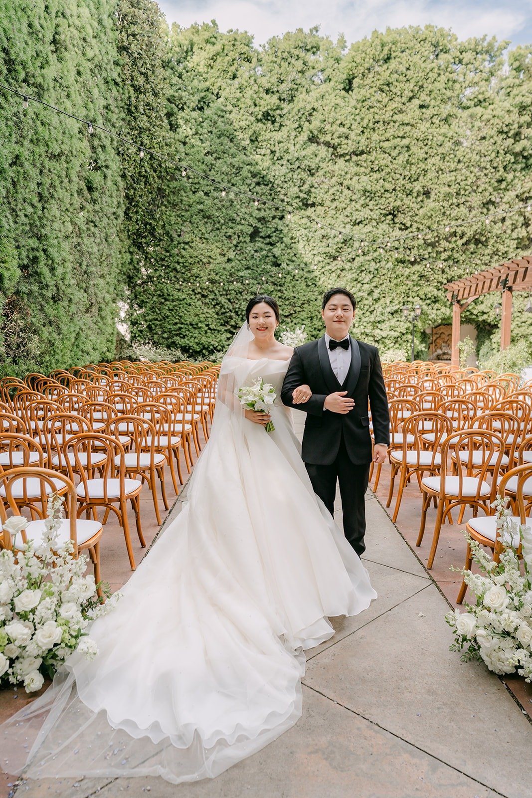 Chorok and Min Soo walking down the empty ceremony aisle between tall green hedges at Franciscan Gardens