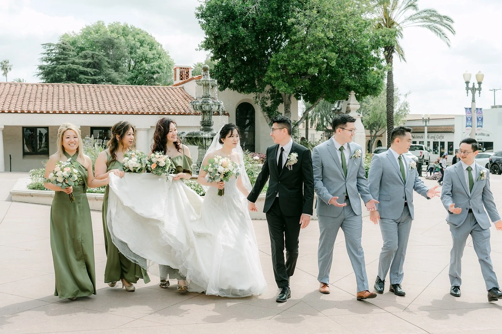 Eunice and Eric's wedding party walking across the plaza at San Gabriel Mission as her veil floats behind her