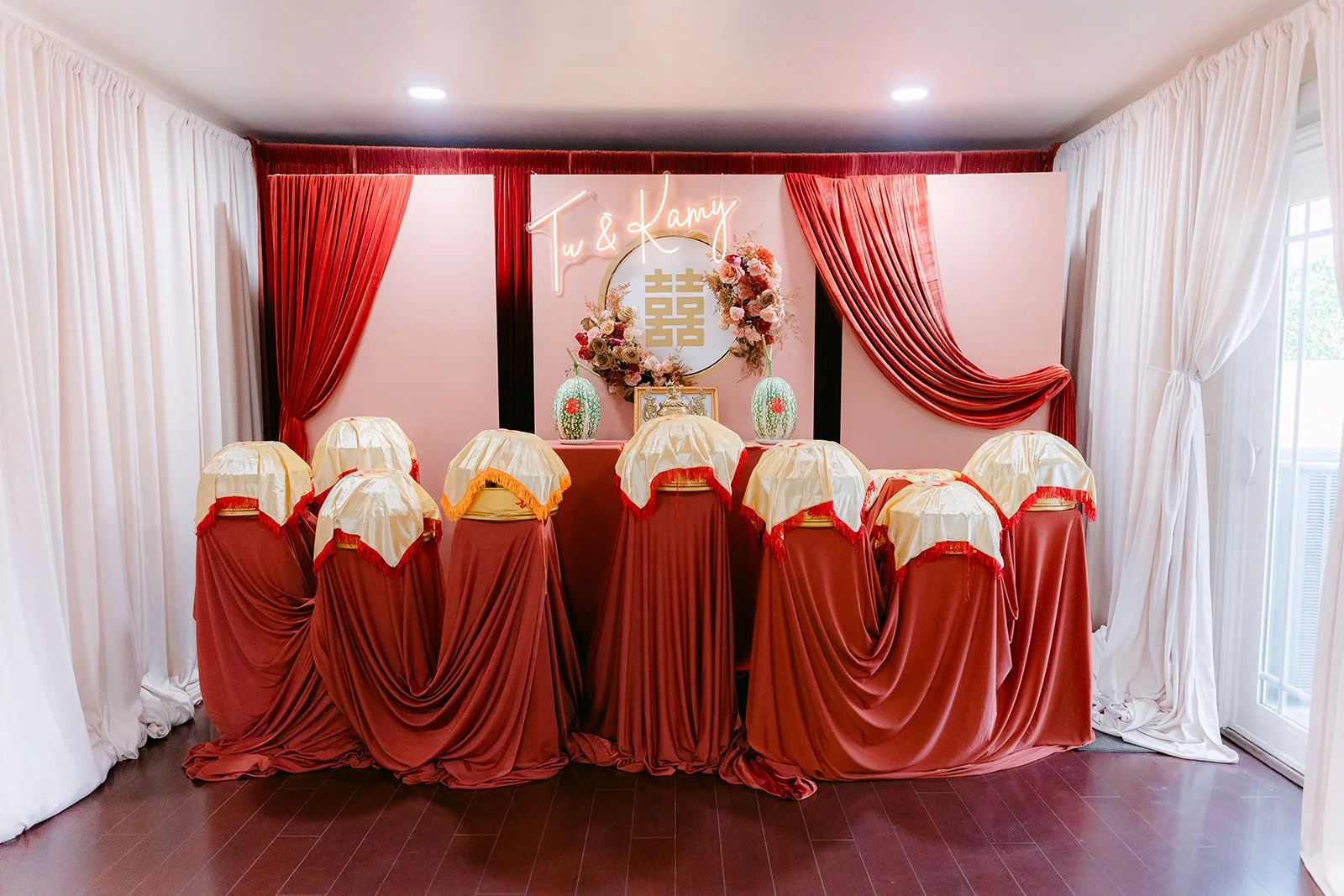 A tea ceremony backdrop with red draped chairs and white curtains set up in a family home.