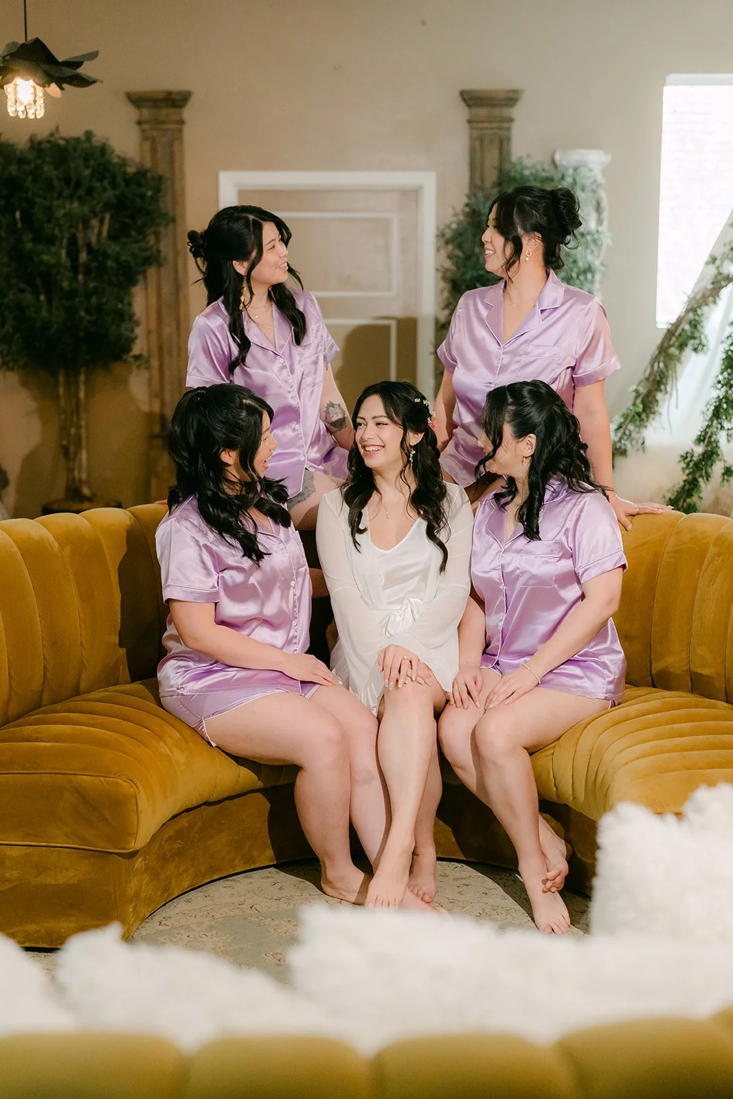 Bride and bridesmaids in lilac pajamas on a mustard velvet sofa