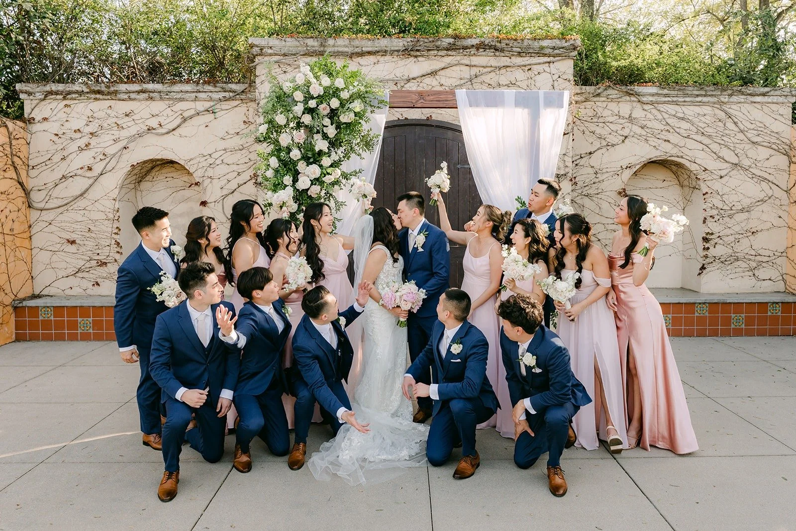 The whole wedding party celebrating in front of the stone wall and floral arch, photographed by One and Only Production