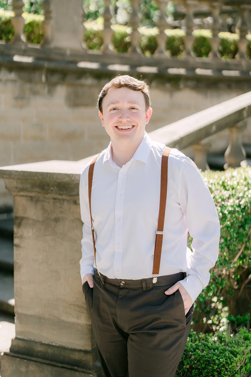 Matthew smiling against the staircase wall in a white shirt and suspenders.