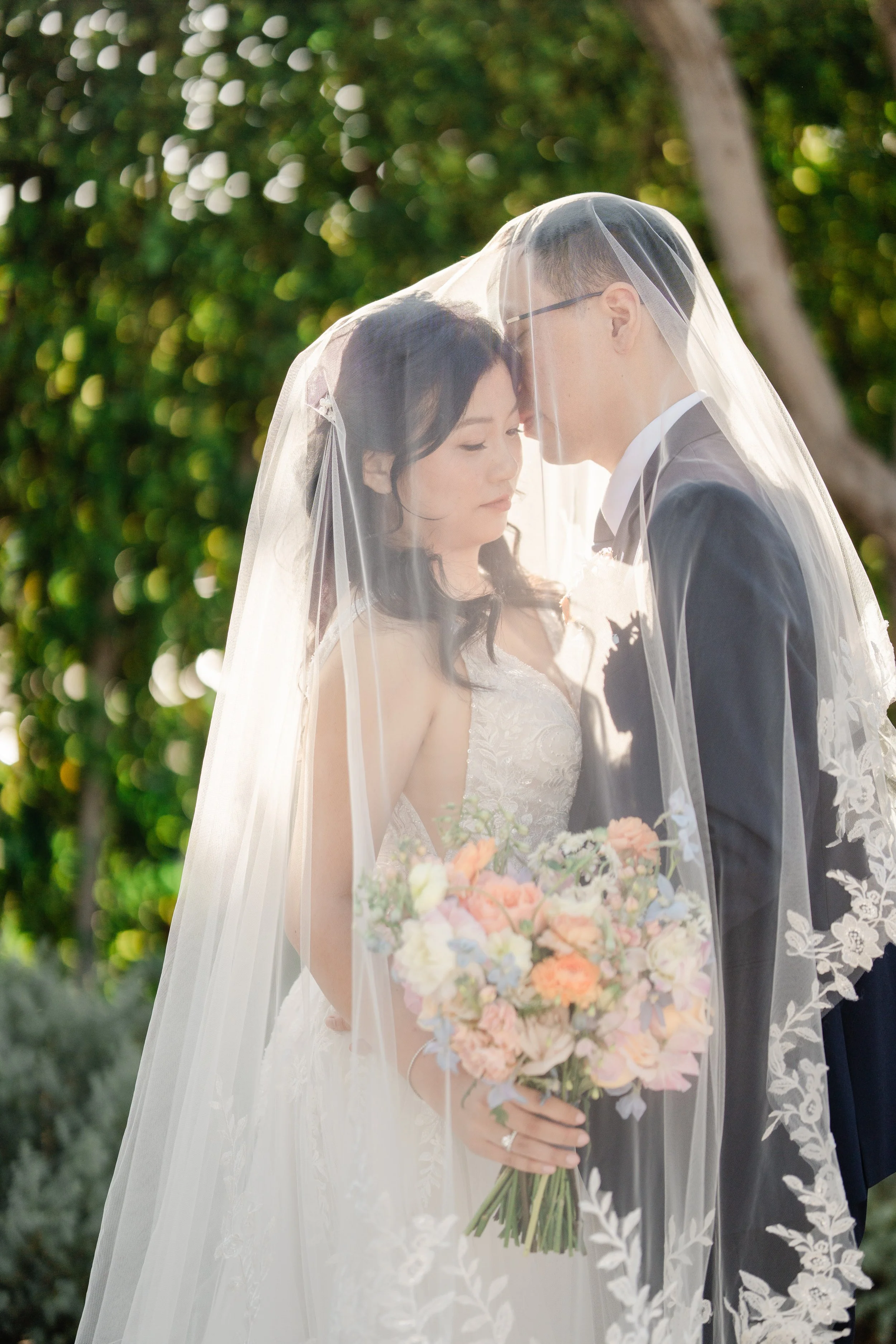 A bride and groom face each other with foreheads touching under a wedding veil, outdoors during daytime, with greenery in the background. The bride holds a bouquet with pastel flowers.