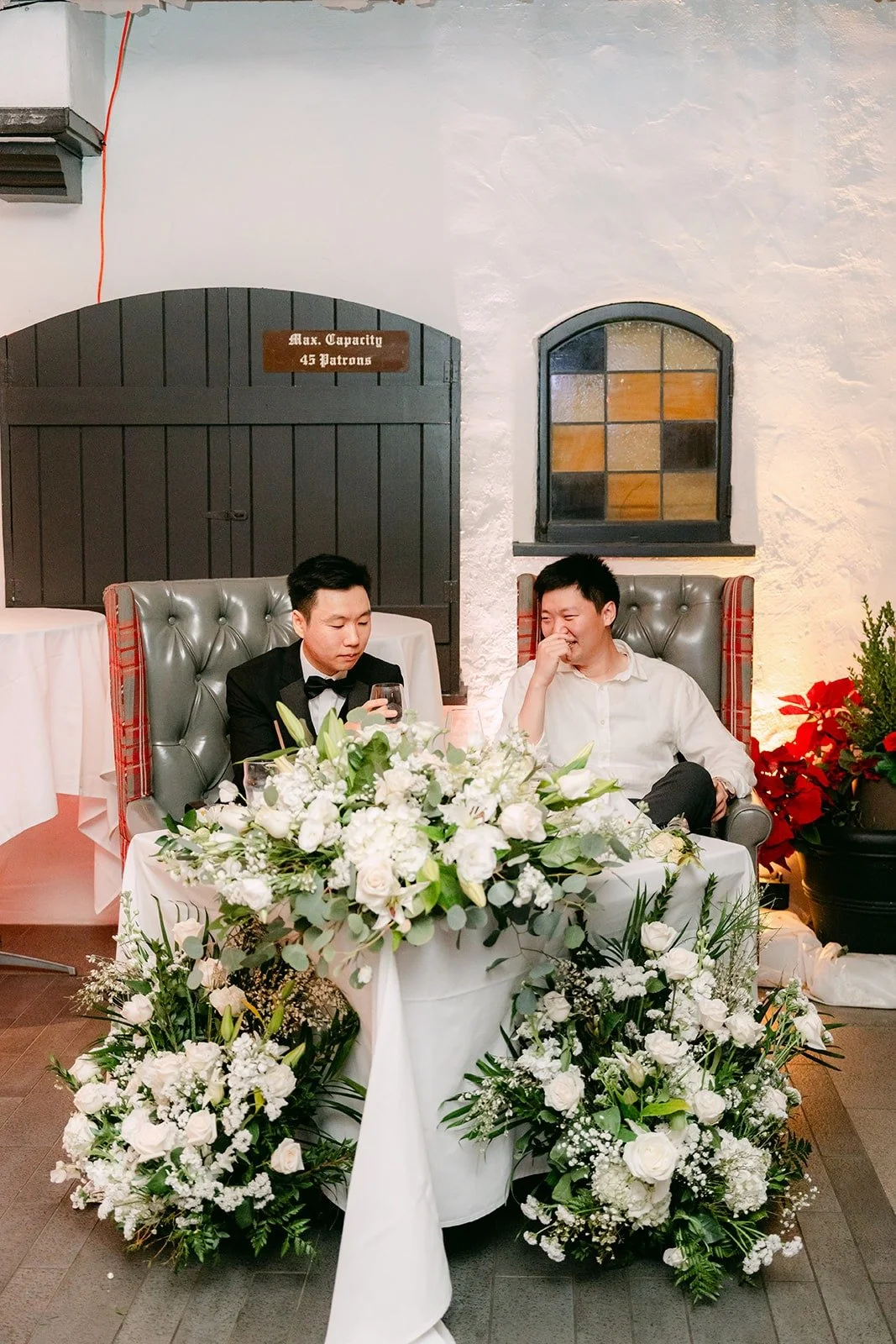 Valerie and Jason at the sweetheart table behind white flowers, photographed by One and Only Production
