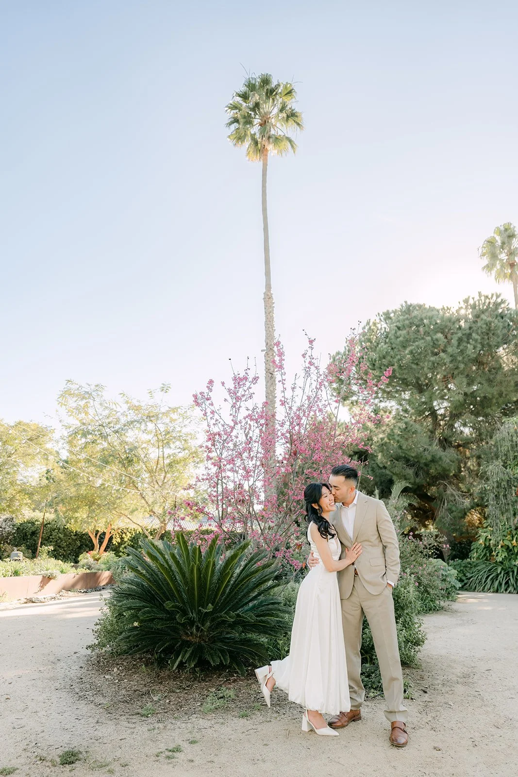Wide view of Kim and Vincent kissing below a tall palm in the garden, photographed by One and Only Production.