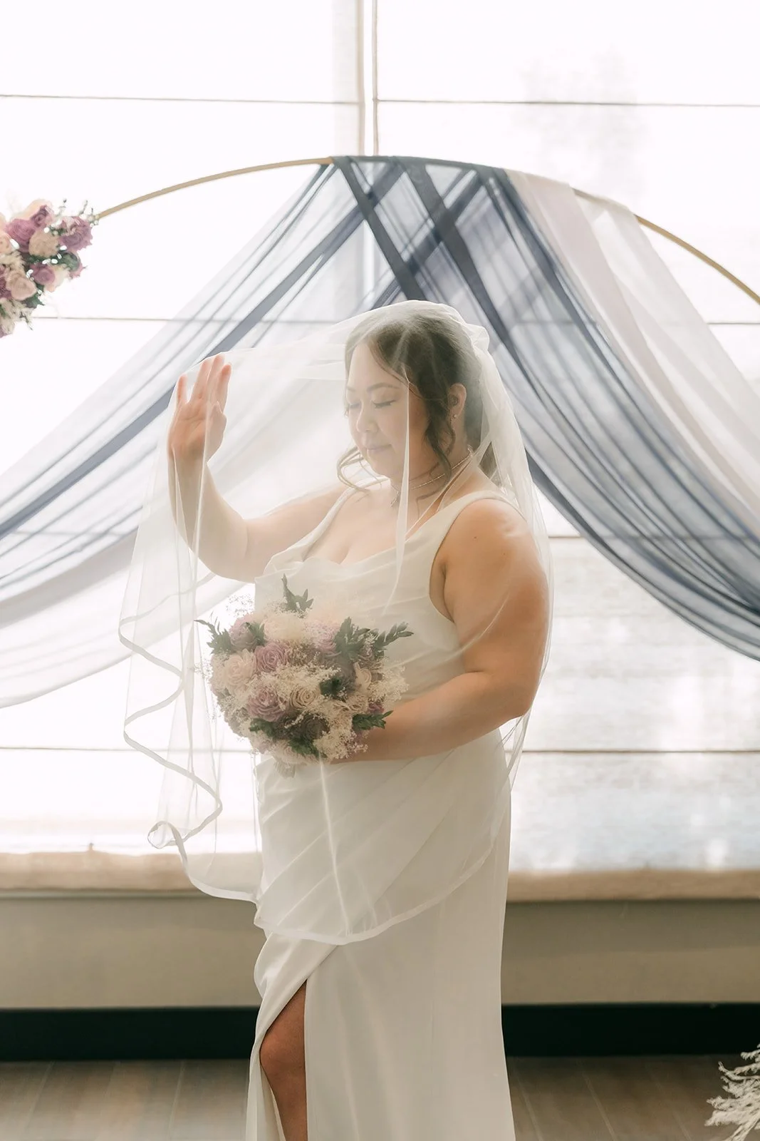 Mai in her gown with her bouquet in front of the draped arch.
