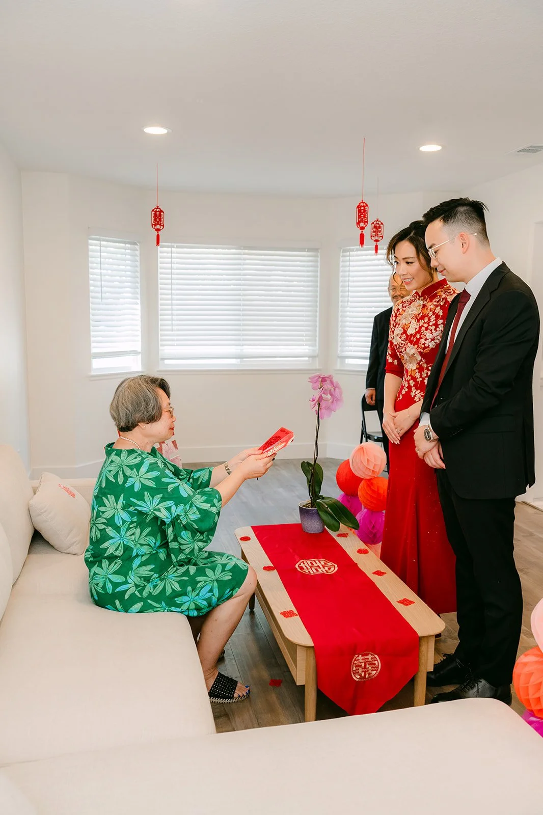 A bride and groom serving tea to an elder seated by the window.